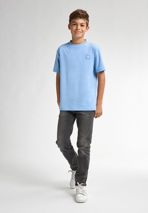 T-shirt di cotone azzurro chiaro con maniche corte, colletto rotondo e piccolo patch logo sul petto sinistro, abbinato a jeans grigio scuro e sneakers bianche.