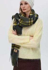Sciarpa a quadri verde, gialla e grigia realizzata in morbida lana, con estremità frangiate e un piccolo etichetta in pelle. Indossata sopra un maglione giallo pallido.
