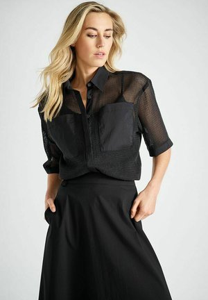 Zwart mesh shirt met korte mouwen, voorzien van twee voorzakken en een knoopsluiting, gecombineerd met een vloeiende zwarte rok.