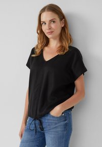 Zwart V-hals top met korte mouwen, trekkoordonderkant en subtiele textuur. Draag met blauwe jeans, benadrukt tegen een neutrale achtergrond.