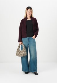 Femme portant un cardigan violet foncé, un haut noir, un jean bleu ample, des chaussures noires, tenant un sac à main beige en simili serpent.