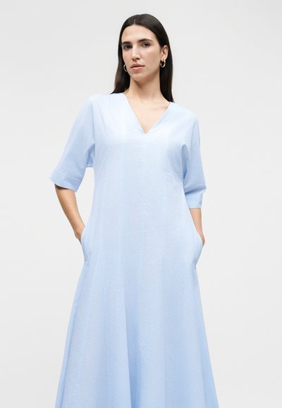Γυναίκα με μακριά σκούρα μαλλιά που φοράει χρυσούς κρίκους σκουλαρικιών και ανοιχτό μπλε φόρεμα με V λαιμόκοψη, μανίκια μέχρι τον αγκώνα, υφή και άνετη εφαρμογή με τσέπες.