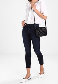 Nainen, joka on pukeutunut valkoiseen paitapuseroon, tummiin slim-fit-farkkuihin, valkoisiin kärkikenkäisiin ja kantaa pientä mustaa kudottua olkalaukkua.