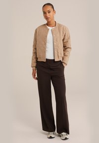 Tan gewatteerde jas met drukknoopsluiting, gedragen over een witte shirt, gecombineerd met bruine wijde broek en grijze sneakers.