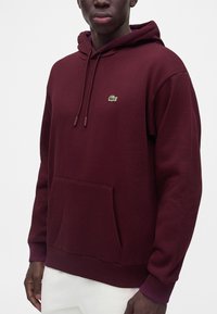 Homme portant un sweat à capuche bordeaux avec une poche avant et un petit logo crocodile vert sur la poitrine, associé à un pantalon blanc.