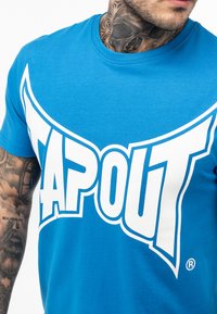 Mėlynas medvilninis marškinėlis su dideliu baltu "TAPOUT" logotipu, su užapvalinta forma ir apvaliu iškirpimu. Matoma rankos detalė su tatuiruotėmis.