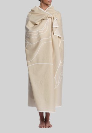 Person, eingewickelt in eine beige Decke mit weißen abstrakten Linien, steht barfuß vor einem schlichten grauen Hintergrund.