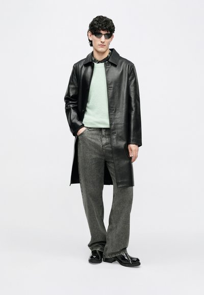 Abrigo largo de cuero negro sobre un suéter de color verde claro, combinado con jeans grises de pierna ancha y botas negras brillantes. Los accesorios incluyen gafas de sol negras.