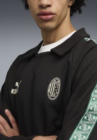 Sort polo shirt med krave, der har grønne mønstrede detaljer på ærmerne, et logo og hvid tekst på brystet. Blød stofstruktur.