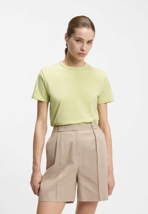 Mujer con el cabello recogido, vistiendo una camisa de manga corta verde claro y pantalones cortos beige de talle alto con rayas, sobre un fondo liso.