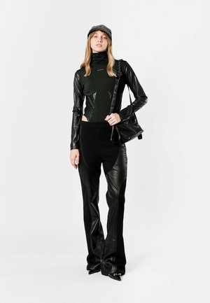 Conjunto negro ajustado que presenta una blusa de cuello alto con acentos de cuero y pantalones acampanados con cortes laterales. Combinado con una gorra y un bolso a juego.