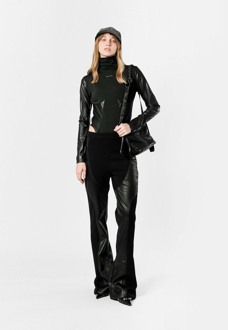 Conjunto negro ajustado que presenta una blusa de cuello alto con acentos de cuero y pantalones acampanados con cortes laterales. Combinado con una gorra y un bolso a juego.