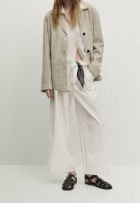 Vaaleanbeige mokkaliivi mustilla napeilla, yhdistettynä vaaleaan paitaan, reisiin asti ulottuviin valkoisiin housuihin ja mustiin kudottuihin sandaaleihin.