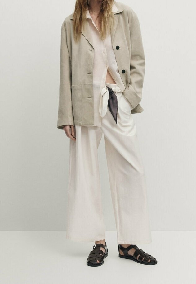 Vaaleanbeige mokkaliivi mustilla napeilla, yhdistettynä vaaleaan paitaan, reisiin asti ulottuviin valkoisiin housuihin ja mustiin kudottuihin sandaaleihin.