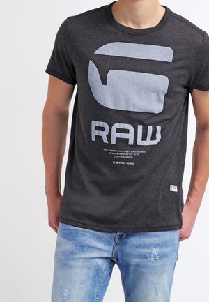 Person iført en mørkegrå G-Star RAW t-shirt med stor lysegrå logo og lyseblå slidte jeans mod en ensfarvet baggrund.