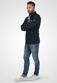 Sudadera azul marino con cordones blancos, bolsillo con cremallera y acentos dorados. Se usa con jeans desgastados azules y zapatillas blancas.