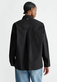Camisa negra de manga larga con cuello estructurado y pliegue en la espalda, con una textura elegante y puños abotonados. Llevada con jeans de un azul claro.