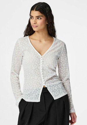 Kobieta z długimi ciemnymi włosami nosi prześwitujący biały sweter w kwiaty na guziki oraz czarne plisowane spodnie z wysokim stanem, patrzy w lewo, z rękami częściowo w kieszeniach.