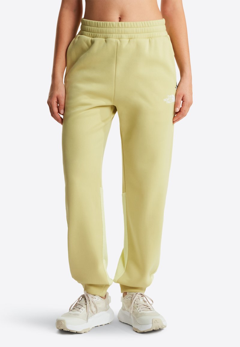 Kobieta w jasnożółtych dresach i beżowych trampkach, stojąca z rękami swobodnie opuszczonymi wzdłuż ciała na białym tle.