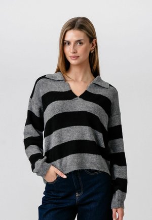 Young woman wearing gray and black striped knit sweater with collar and blue jeans, standing with one hand in pocket against plain background.