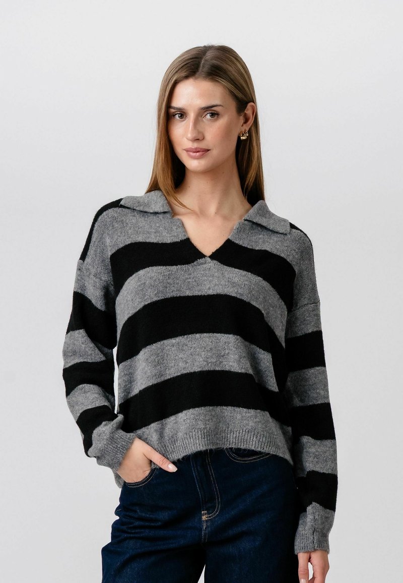 Ung kvinde iført grå og sortstribet strikket sweater med krave og blå jeans, stående med den ene hånd i lommen mod en ensfarvet baggrund.