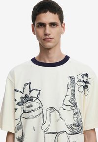 Cream cotton T-shirt with a navy crew neckline, featuring black line-drawn illustrations of a vase, teapot, and flowers.