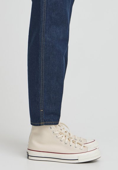 Baskets montantes en toile beige avec semelles en caoutchouc blanches, accent en stripe rouge, associées à un jean en denim bleu foncé avec des coutures visibles.