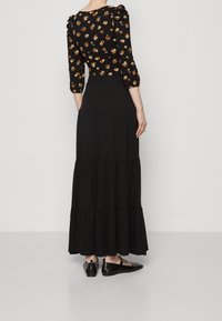 Kvinna klädd i en svart, lager-på-lager maxi kjol, svart blommig långärmad topp med gula och orange blommor, och svarta Mary Jane-skor, sedd bakifrån.