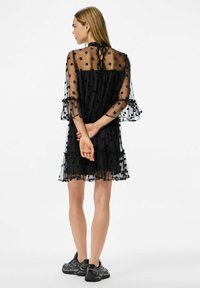 Crna haljina od prozirnog materijala s točkicama, naboranim rubovima i rukavima u obliku zvona. Ima mašnu na vratu. Nosena s crnim tenisicama.