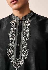 Black shirt featuring intricate white embroidery along the front placket, with a stand collar and decorative buttons. Smooth fabric texture.