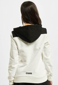 Sudadera blanca con detalles negros en los hombros y la capucha. Presenta puños y dobladillo acanalados, textura suave, con el logo "DNGRS" en la parte baja de la espalda.