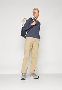 Hanorac bleumarin cu un logo, pantaloni bej și încălțăminte de drumeție multicolore. Persoana poartă un rucsac negru și gri cu un capac în carouri.