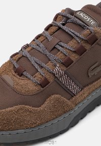 Braunfarbener Sneaker mit texturiertem Wildleder- und Mesh-Obermaterial, grauer Gummisohle und detaillierter grauer Schnürung. Sichtbares Lacoste-Logo.
