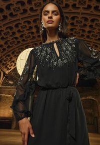 Rochie neagră cu mâneci translucide, volanate, detalii brodate cu stele și lună argintii și un decolteu în formă de lacăt cu o panglică la talie.