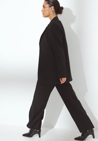 Črn krojen suit z večjim blazerjem in ravnimi hlačami, v kombinaciji s črnimi čevlji s peto, postavljenimi na belo ozadje.