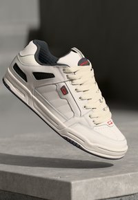 Cream-colored sneaker with a textured finish, featuring grey accents, a lace-up design, and a black padded collar. Notable red logo detail.