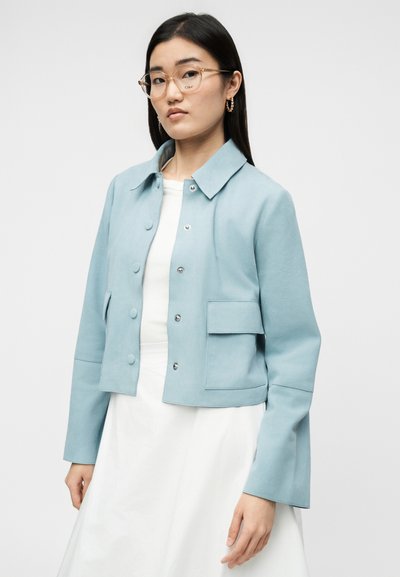 Γυναίκα που φοράει γαλάζιο σακάκι με κουμπιά και τσέπη με καπάκι πάνω από λευκό φόρεμα, μπεζ γυαλιά και χρυσούς κρίκους στα αυτιά, στέκεται μπροστά σε λευκό φόντο.