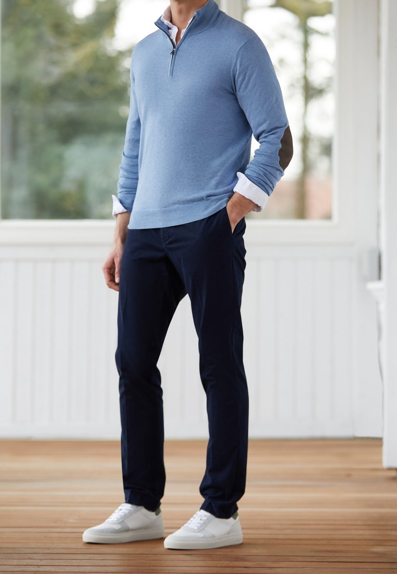 Ljusblå kvartdraget pullover med vit krage och bruna axelaccenter, tillsammans med marinblå byxor och vita sneakers med gröna detaljer.