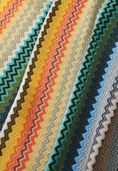 Cobertor colorido tecido à mão com padrões em zigue-zague nas cores laranja, amarelo, verde, azul e bege, em um tecido texturizado com um acabamento suave.