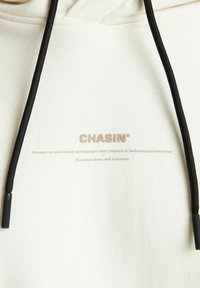 Crème hoodie met zwarte trekkoorden. Bevat een logo "CHASIN'" en tekst afgedrukt in lichtgrijs. Zachte stof met een gladde textuur.