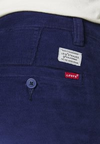 Námořnické kordové kalhoty s modrým knoflíkem, na nichž je bílá etiketa s nápisem "Levi Strauss of California" a červený štítek "Levi's".