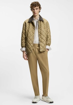 Veste matelassée kaki avec un col marron, associée à un pantalon beige et des baskets blanches. La veste est dotée de boutons-pression et d'un détail logo.