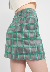 Mini-jupe à carreaux verts et noirs en tissu texturé, présentant une coupe ajustée et des poches latérales. Motif géométrique clair.