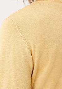 Zbliżenie na ramię żółtego swetra z dzianiny z widocznym szwem i teksturą, na osobie z jasno brązowymi włosami częściowo widoczną.