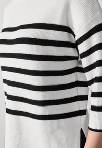 Pullover rayé en blanc et noir, présentant des rayures horizontales, une texture côtelée et des fentes sur les côtés. Fabriqué en matériau tricoté doux.