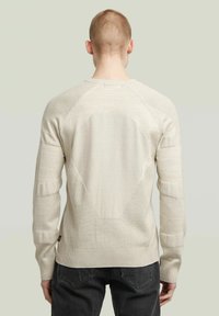 Strickpullover in hellem Beige mit strukturierten Einsätzen, gerippten Bündchen und einem markanten Rautenmuster auf der Rückseite, kombiniert mit dunklen Jeans.