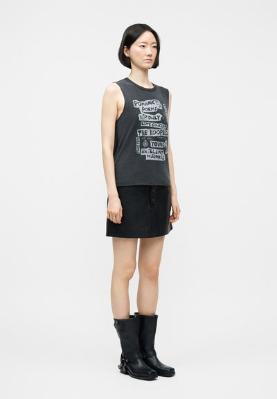 Ασιατίδα γυναίκα που στέκεται, φοράει μαύρο αμάνικο τοπ με γραφικό σχέδιο και κείμενο, μαύρη μίνι φούστα και μαύρες μπότες μέχρι τη μέση της γάμπας σε απλό φόντο.