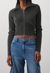 Beskåret, mørkegrå jakke med lynlås, højt krave og lange ærmer, parret med lyseblå denim cargo bukser med sidelommer.