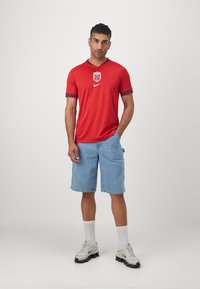 Maglia sportiva rossa con uno stemma blu e bianco, abbinata a pantaloni corti di denim azzurri e scarpe sportive grigie con suole nere.
