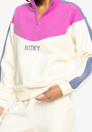 Mujer con un pulóver colorblock con mangas rosas, crema y azules, con una cremallera a medio subir y el logo "ROXY" en el pecho.